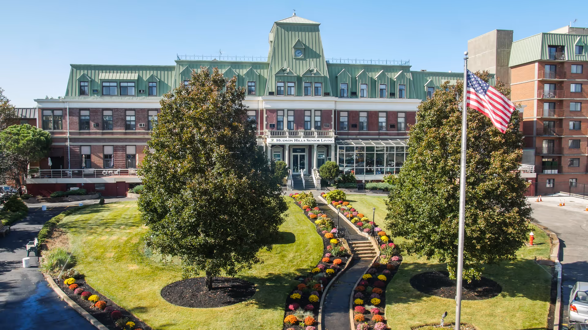 Front exterior of a senior living facility showing the building facade, landscaped lawn and walkway, trees, and an American flag.