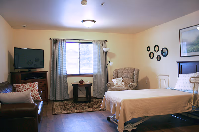 Furnished assisted living bedroom with a hospital-style bed, armchair, sofa, TV, and a window with curtains.