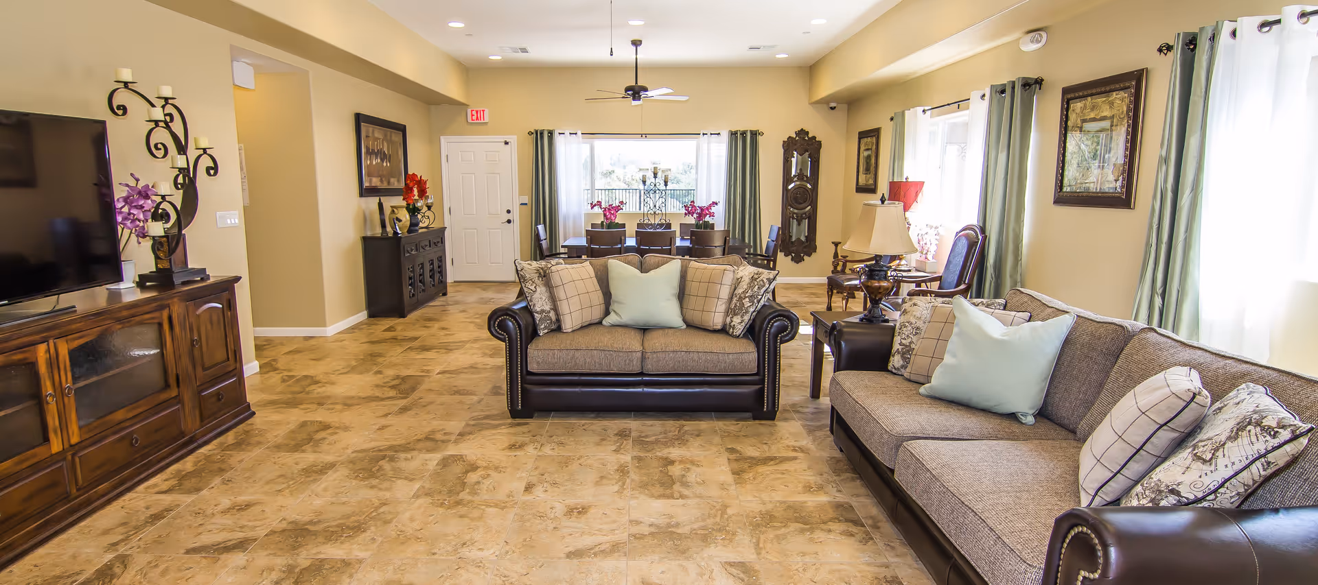 A spacious living room with two sofas adorned with multiple pillows, a large flat-screen TV on a wooden cabinet, a dining table with chairs near a window, decorative lamps, framed artwork on the walls, and large windows with curtains allowing natural light to fill the room.