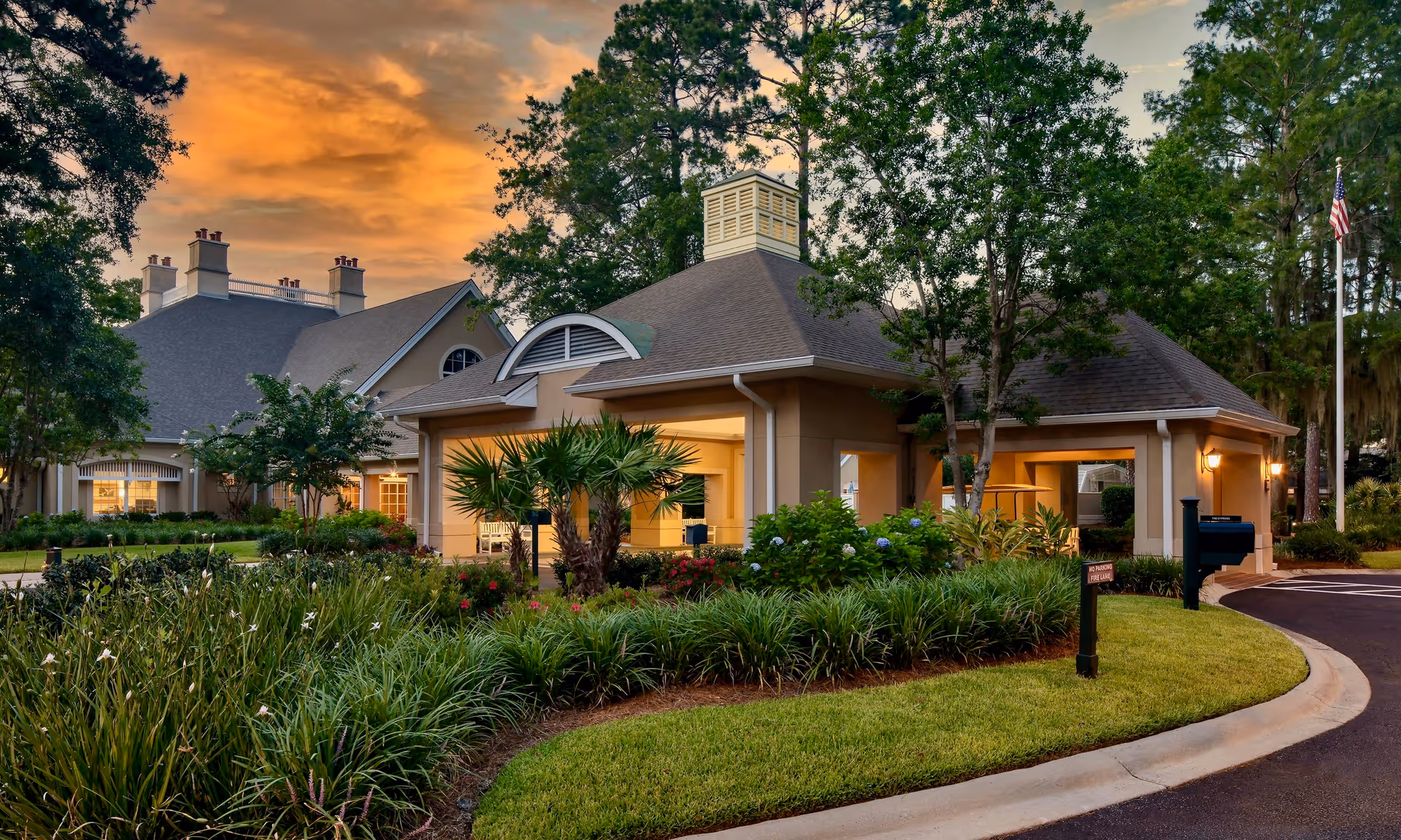 Exterior view of a senior living facility building at sunset with well-maintained landscaping including green shrubs, palm trees, and flowering plants. The building has a beige facade with a gray roof and multiple chimneys. There is a curved driveway and an American flag on a flagpole to the right.