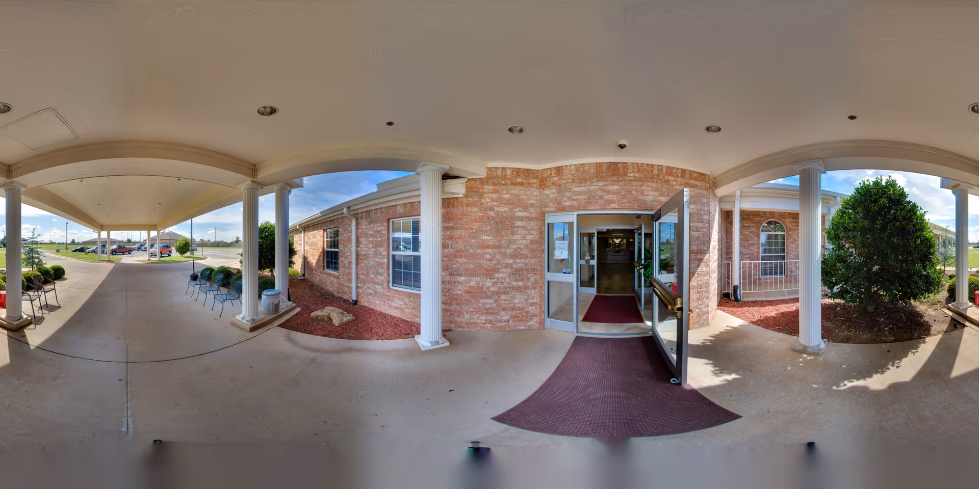 Covered entrance area of a brick building with white columns and a glass door open to the interior. There are chairs lined up along the left side under the covered walkway, and landscaping with bushes and mulch on the right side. The sky is partly cloudy and the parking lot is visible in the background.