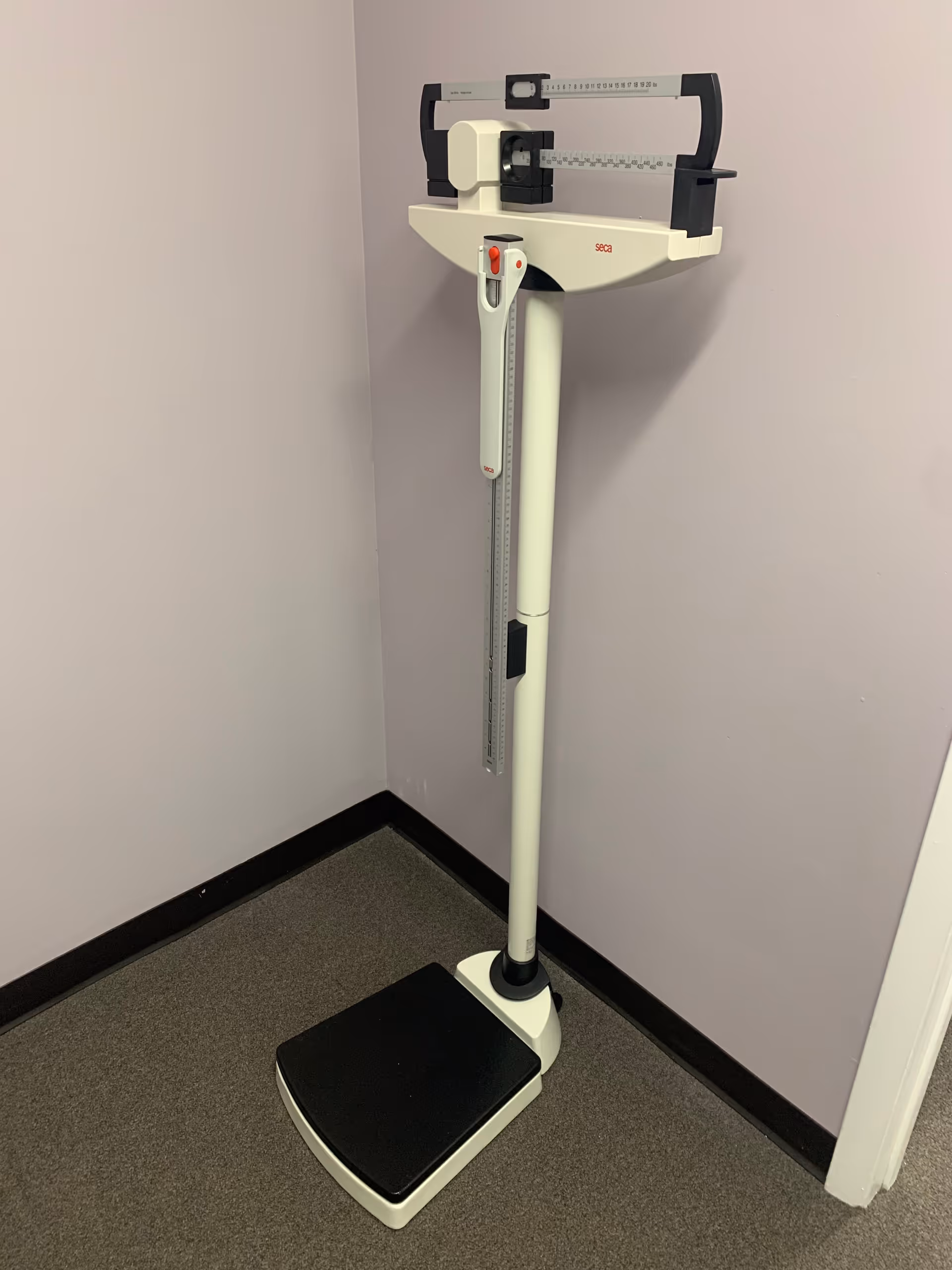 A standing medical scale with height rod in a corner of a small clinical room.