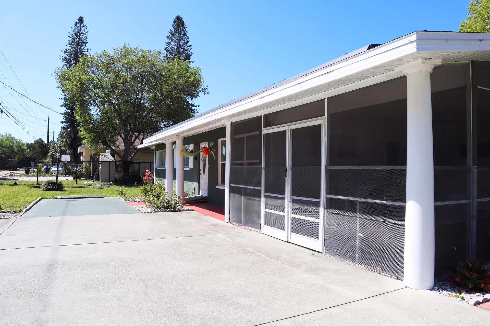 Exterior view of a single-story building with a screened porch supported by white columns. There is a concrete driveway and a small garden area with a tree and some plants. The sky is clear and blue.