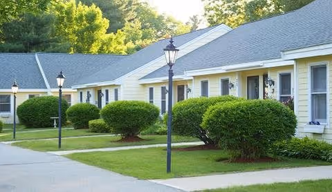 Row of single-story residential units with white siding and gray roofs, each with a small porch light and neatly trimmed bushes in front. A paved walkway runs alongside the units with black lamp posts lining the path. Trees and greenery are visible in the background under a clear sky.
