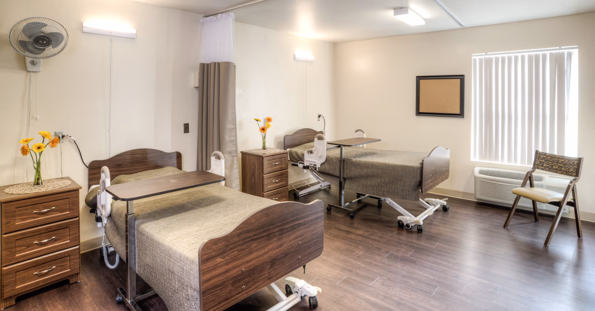 A bright shared patient bedroom with two adjustable hospital beds, over-bed tables, dressers, chairs and a curtain divider.
