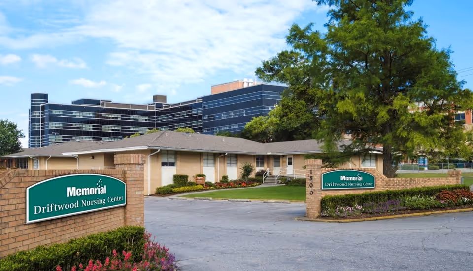 Exterior view of Memorial Driftwood Nursing Center showing a single-story brick building with a driveway and landscaped greenery including bushes and flowers. Two green signs with white text display the facility name on brick pillars at the entrance. A large tree is visible on the right side, and a multi-story modern building is in the background under a partly cloudy sky.