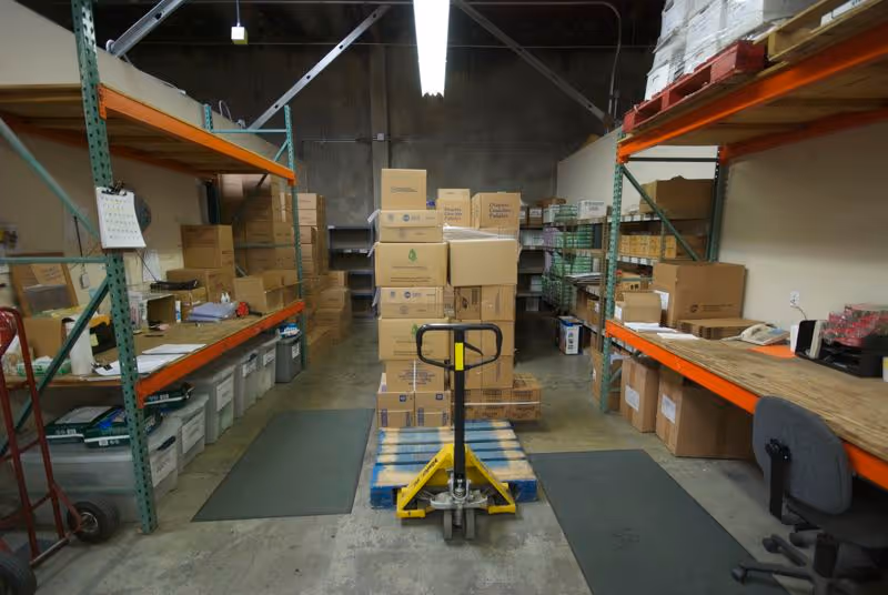 Interior storage room full of boxes, shelves, a pallet jack, and a desk.