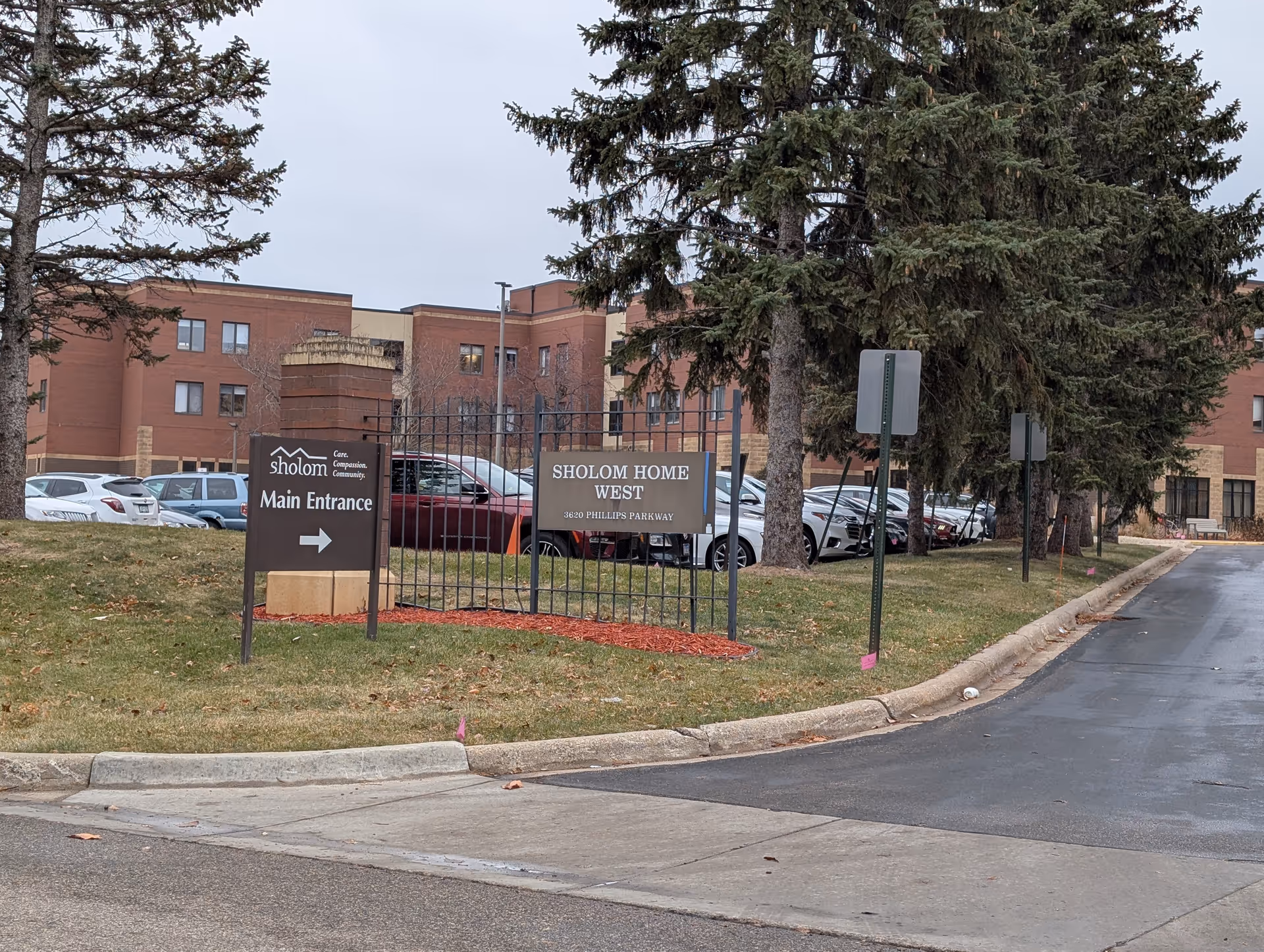Entrance signs for 'Sholom Home West' and 'Main Entrance' beside a driveway leading to a brick assisted living building with parked cars and trees.