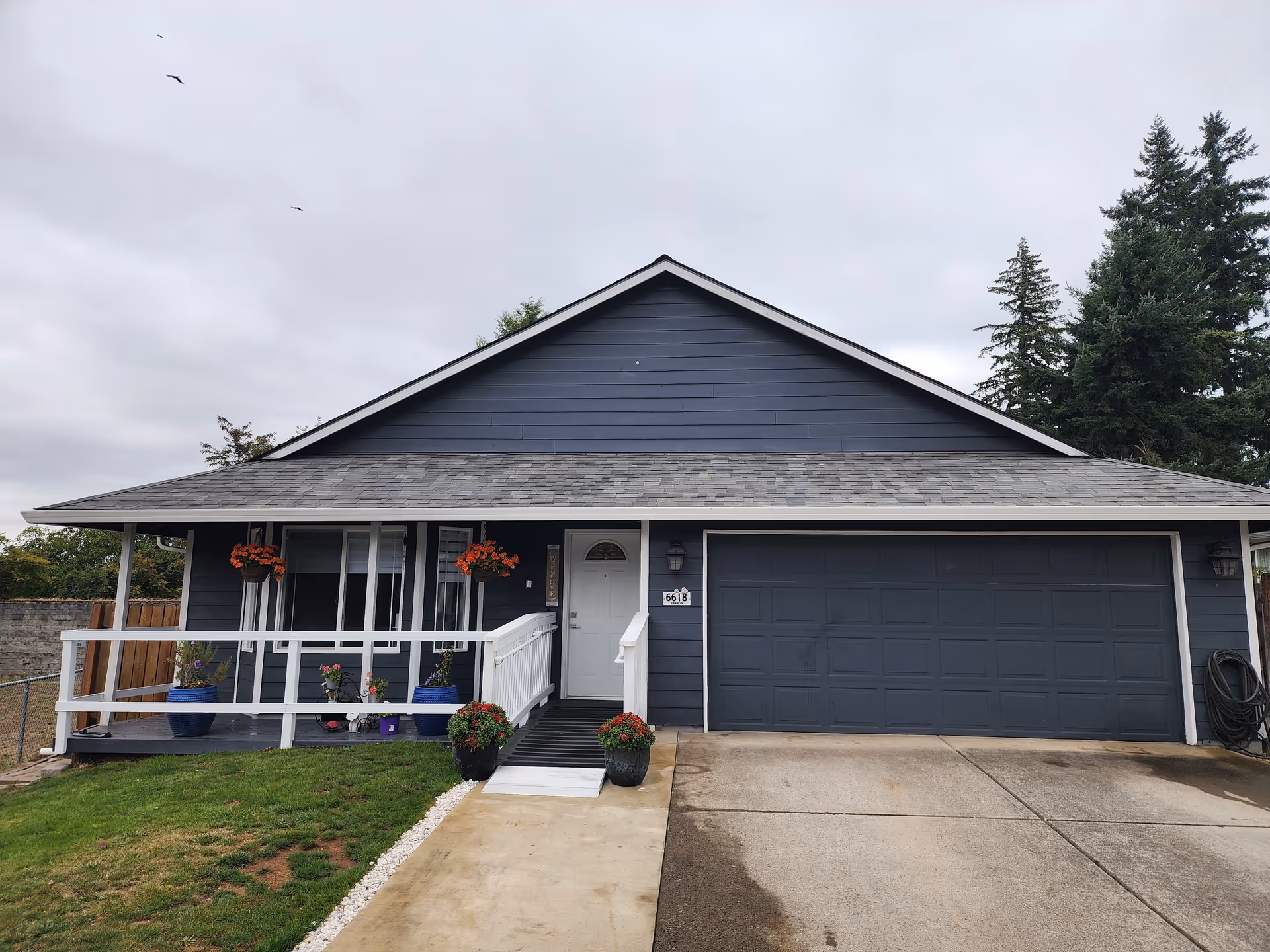 Blue-gray single-story house with a front porch, white railing, potted plants, and a two-car garage.