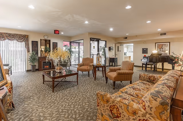 Cozy senior living lounge with patterned sofas, chairs, coffee table, piano, and fireplace.