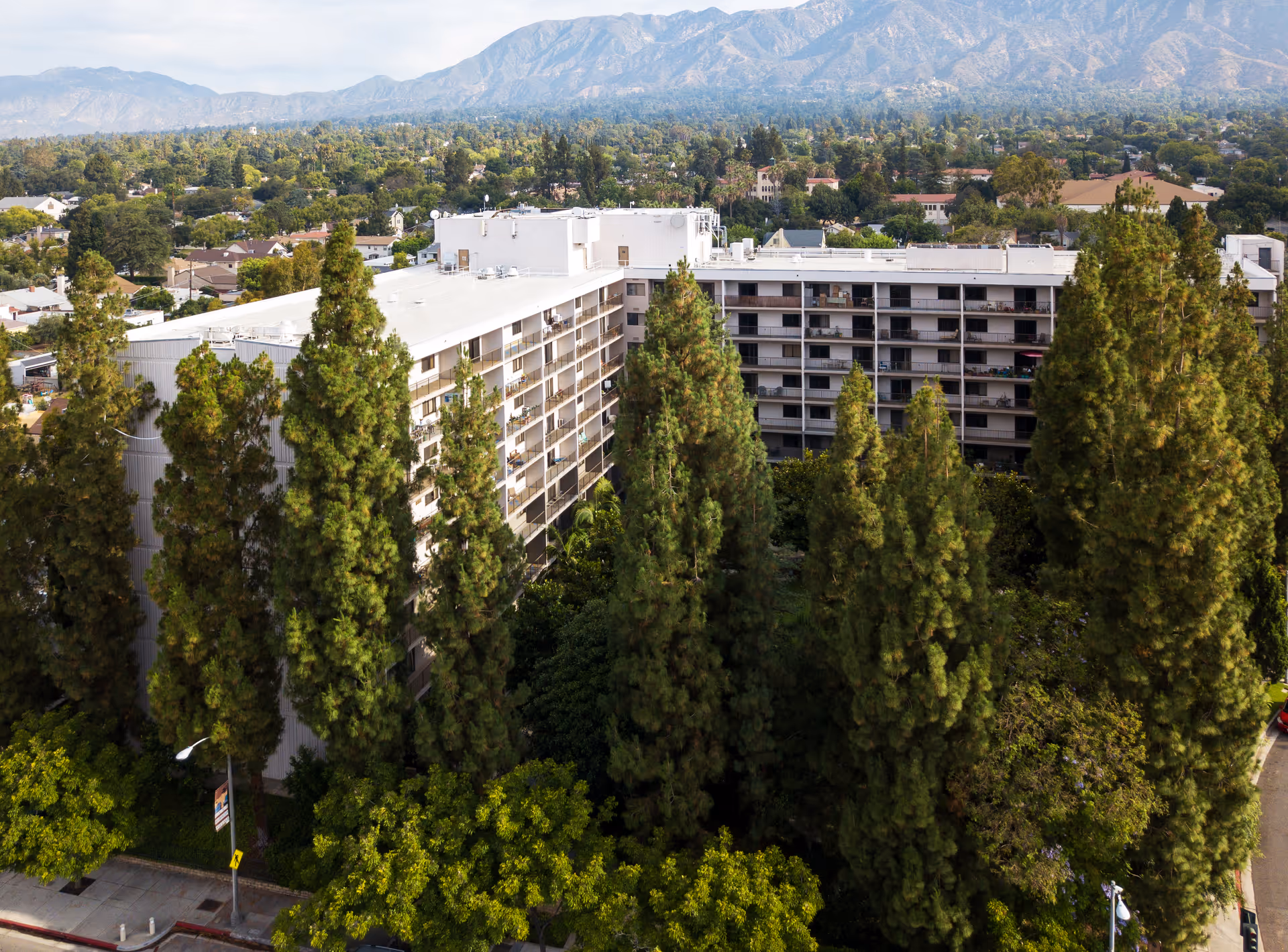 Aerial view of a multi-story residential building surrounded by tall green trees with a mountainous landscape in the background under a partly cloudy sky.