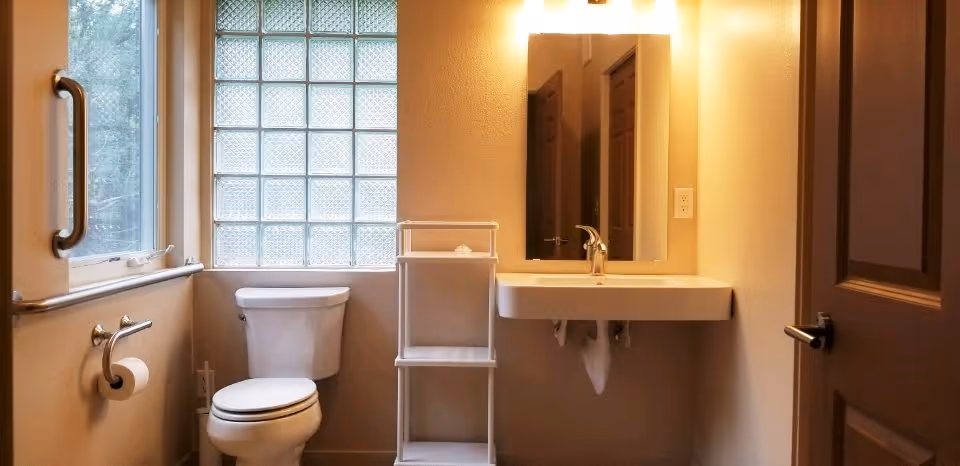 A small bathroom with a white toilet, a wall-mounted sink with a faucet, a rectangular mirror above the sink, a white shelving unit between the toilet and sink, a window with frosted glass blocks, and a grab bar on the wall next to the toilet. The walls are painted beige and the door is partially open.
