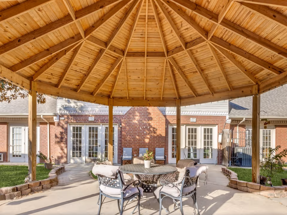 Outdoor seating area under a wooden gazebo with a round metal table and four cushioned chairs. The background shows a brick building with multiple glass doors and windows, and some greenery around the patio.