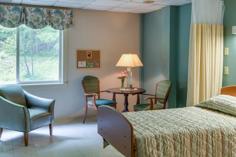 A tidy senior facility bedroom with a single bed, two chairs, a small round table with a lamp and flowers, and a window showing greenery.