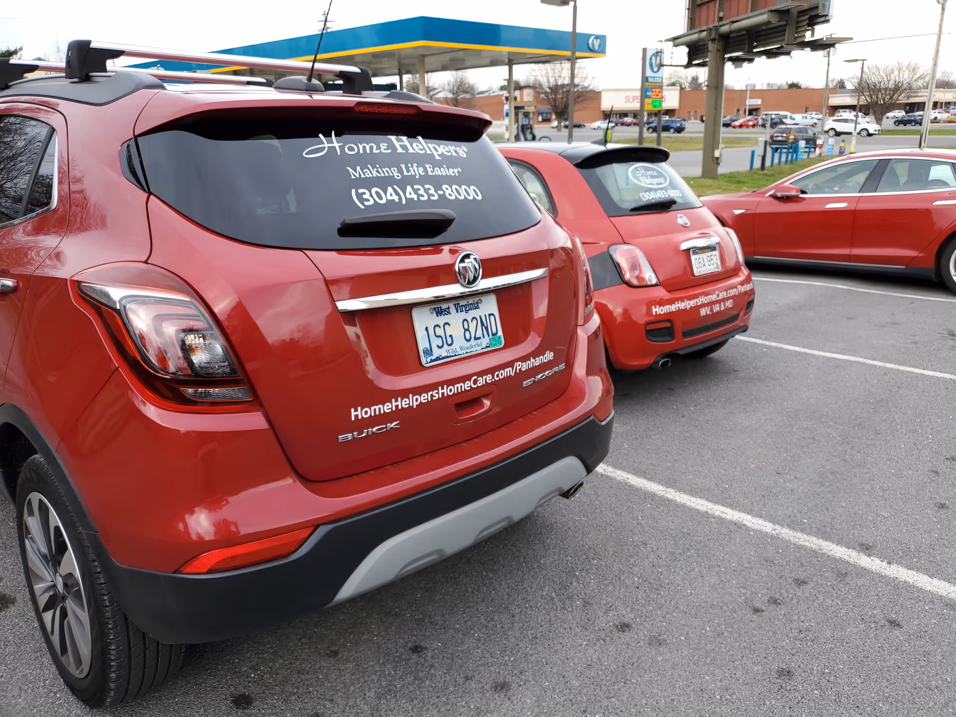 Red branded Home Helpers vehicles parked in a lot by a gas station.