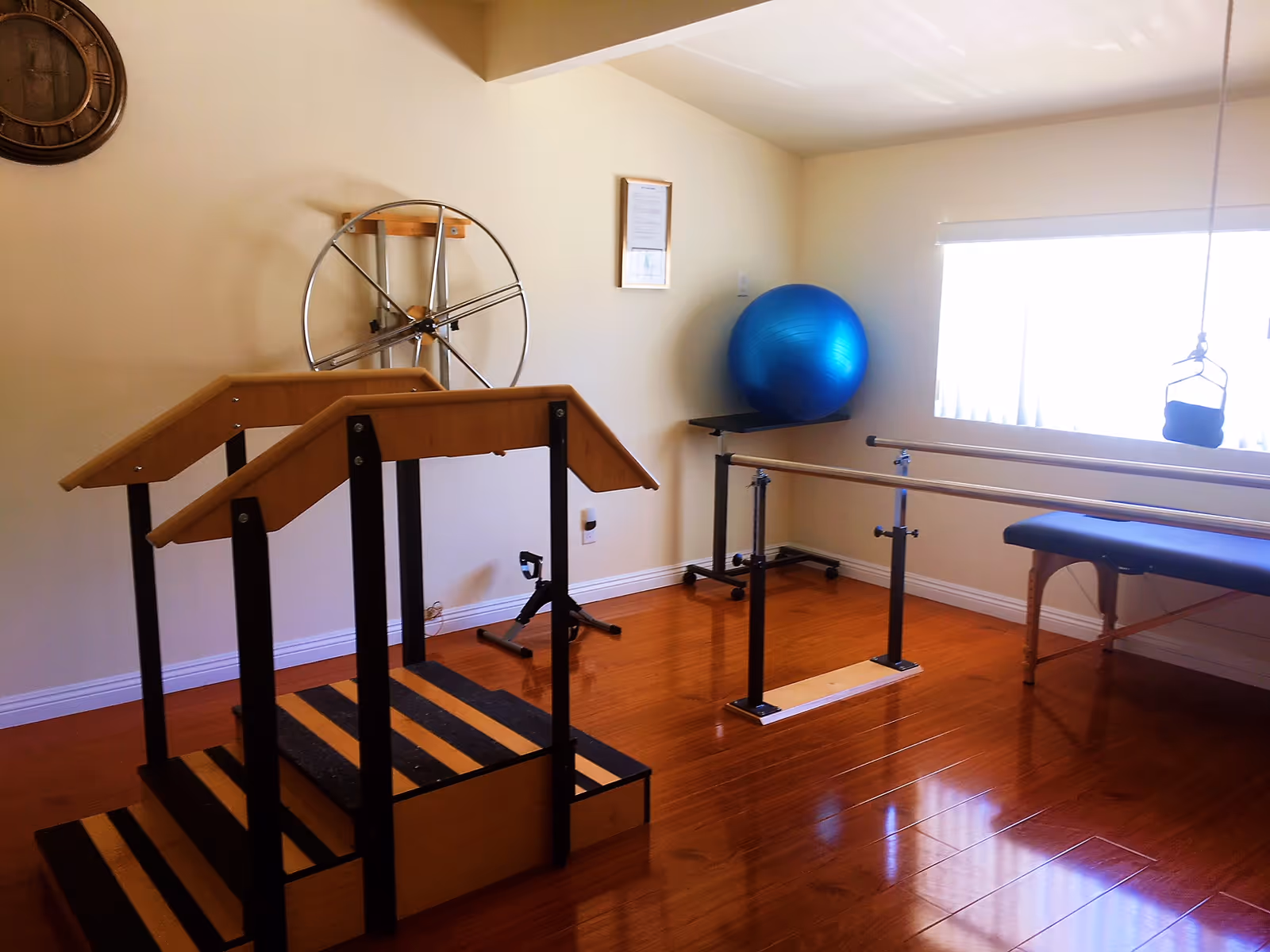Interior rehab exercise room with parallel bars, a large wheel device, blue stability ball, and window.