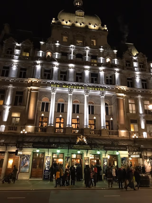Nighttime exterior of a grand theatre with illuminated facade and a crowd outside.