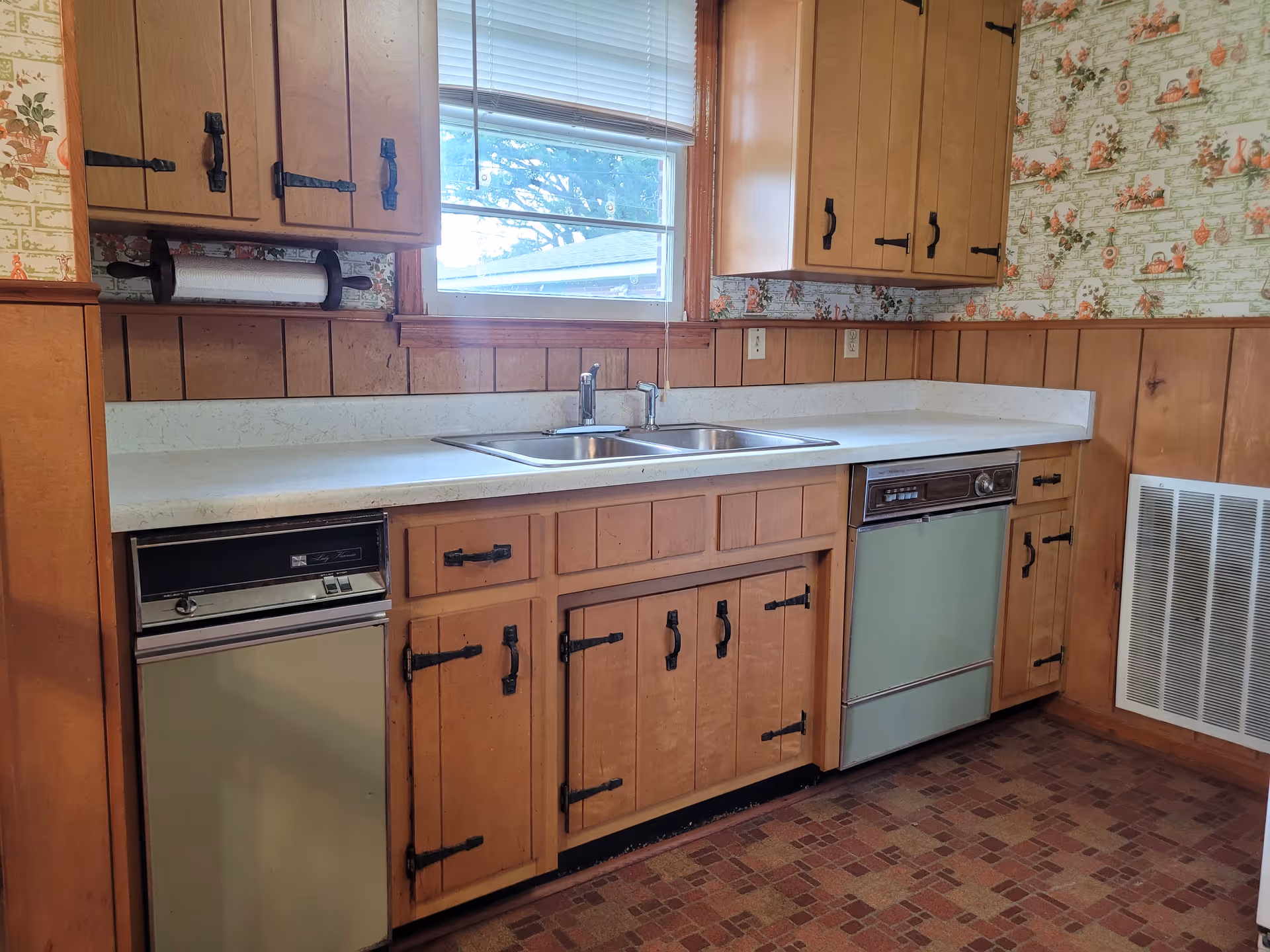 Wood-paneled kitchen with sink, dishwasher, stove, and vintage wallpaper behind cabinets.