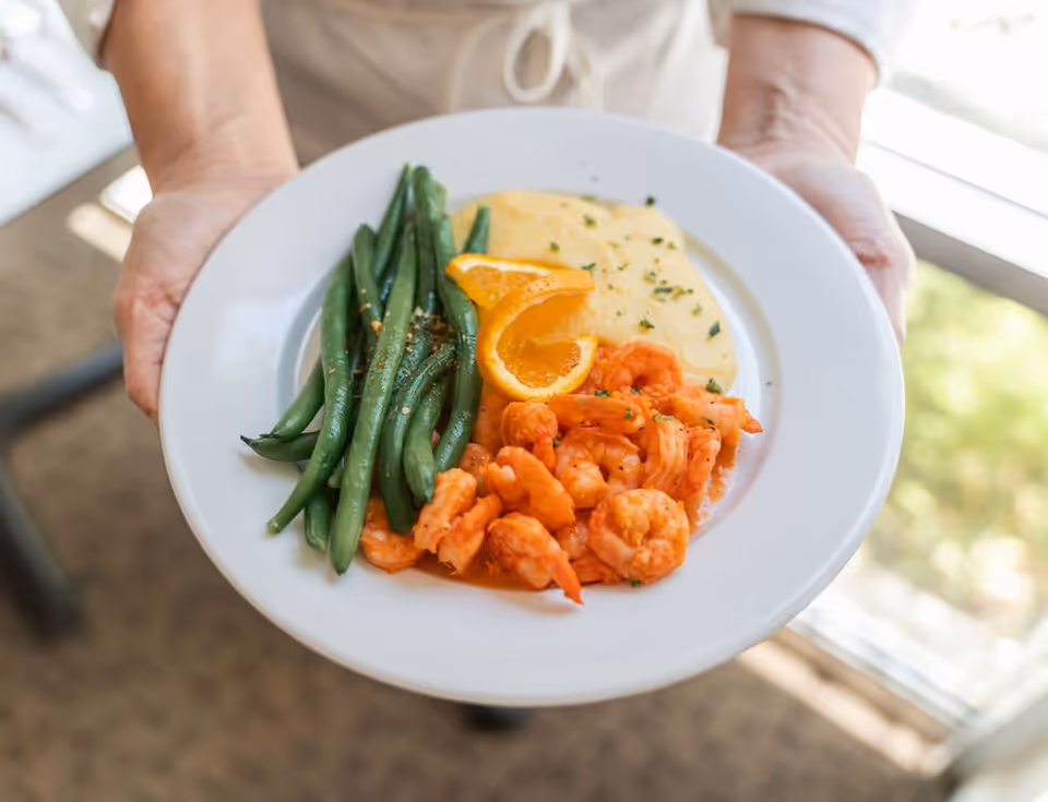 A person holding a white plate with a meal consisting of cooked green beans, shrimp, a serving of creamy yellow grits, and a garnish of orange slices.