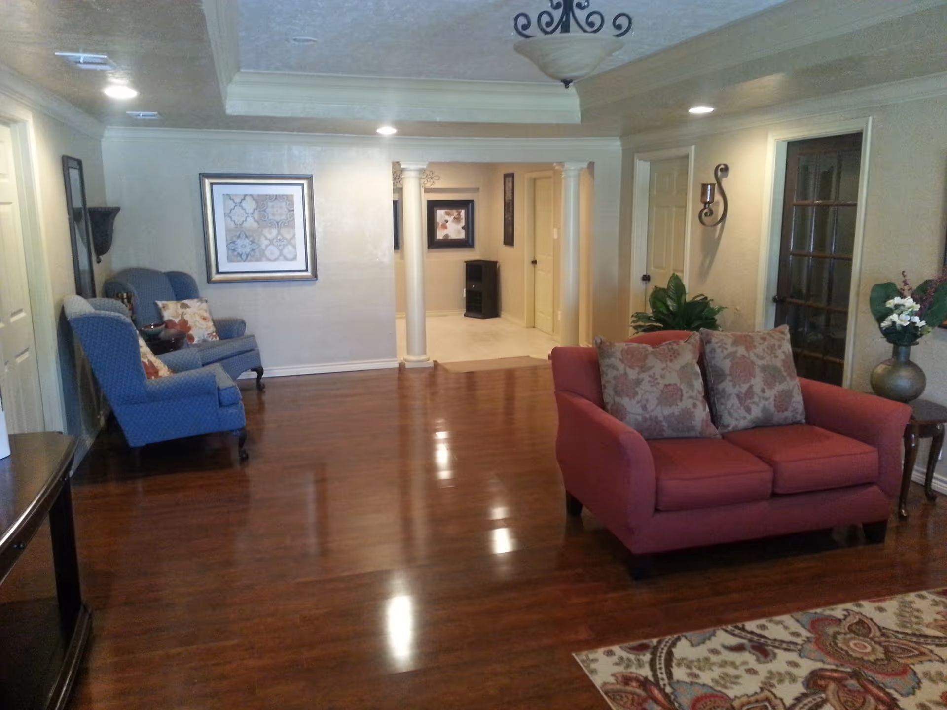 Spacious living room with polished hardwood floors, a red loveseat, blue armchairs, decorative pillows, wall art and columns.