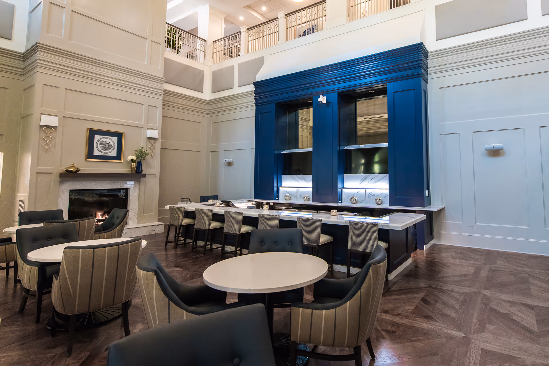 A spacious and elegant common area with high ceilings, featuring a fireplace on the left wall, several round tables with cushioned chairs, and a large blue built-in unit with multiple cooking or warming stations. The room has wood flooring and light-colored paneled walls with modern wall sconces.