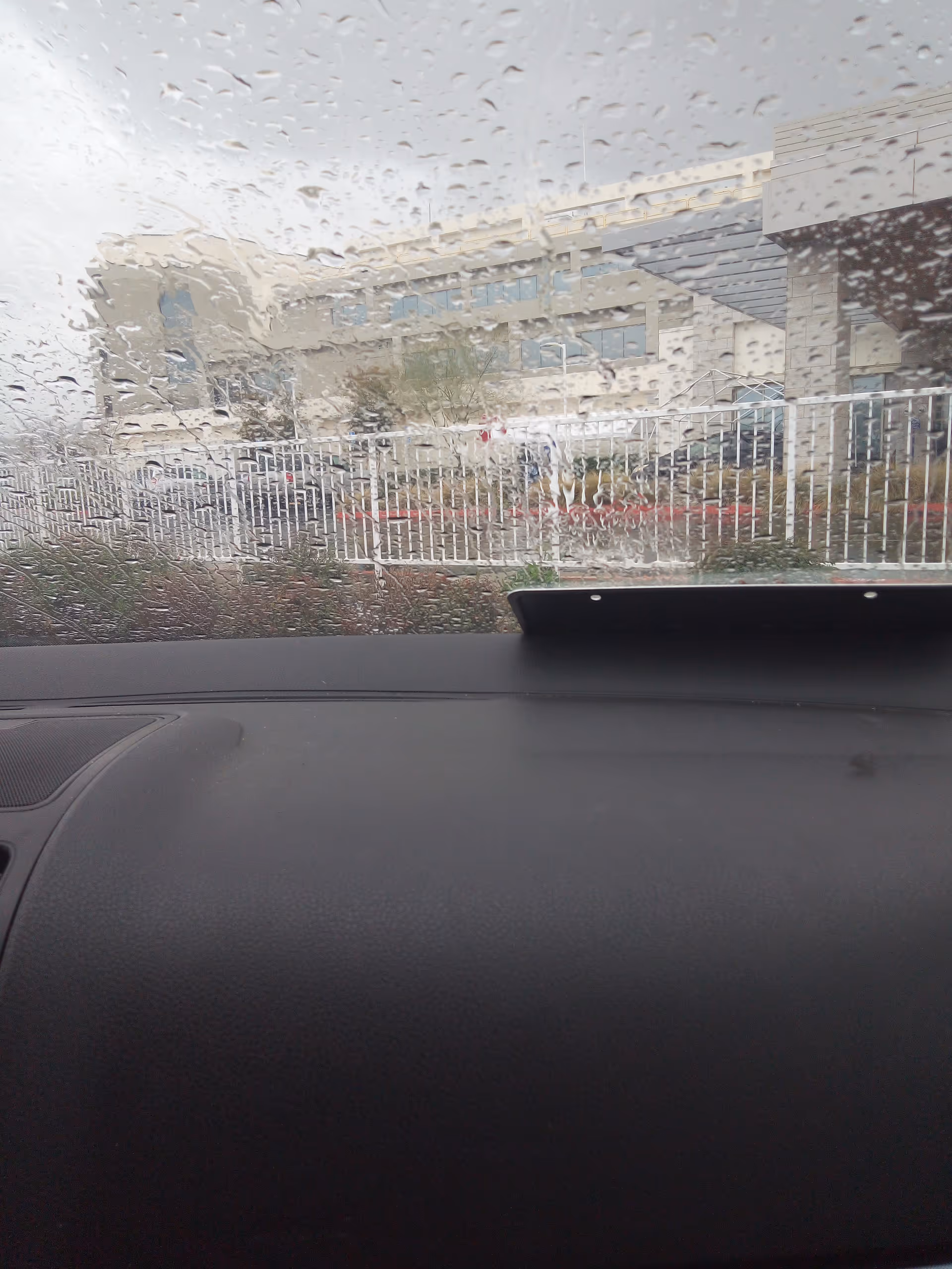 Rainy windshield view of a modern facility exterior behind a white fence