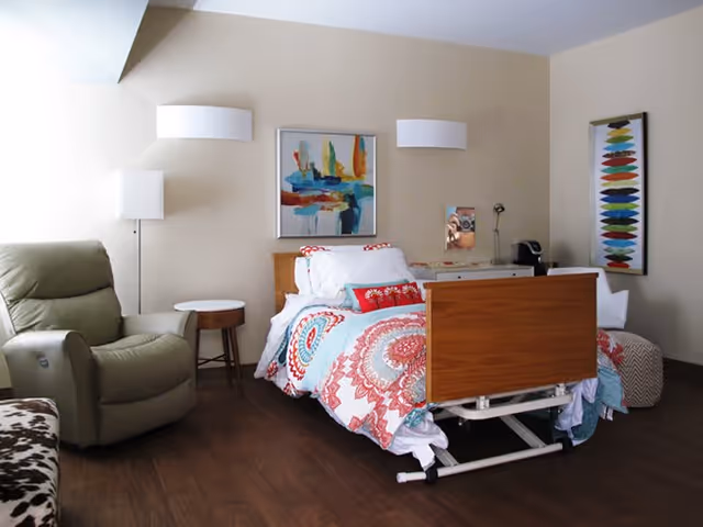 A furnished bedroom with an adjustable hospital-style bed covered in colorful bedding, a recliner, side tables and wall art.