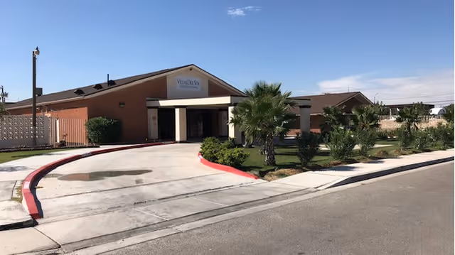 Front entrance of the Villas Del Sol assisted living building with a covered drop-off, circular driveway, and palm landscaping under a clear sky.