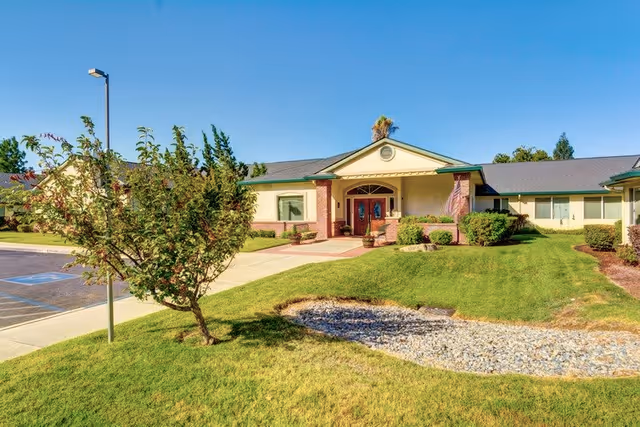 Front exterior view of Cogir of Turlock facility showing a single-story building with a covered entrance, double wooden doors, well-maintained green lawn, small trees, shrubs, and a dry creek bed with rocks under a clear blue sky.