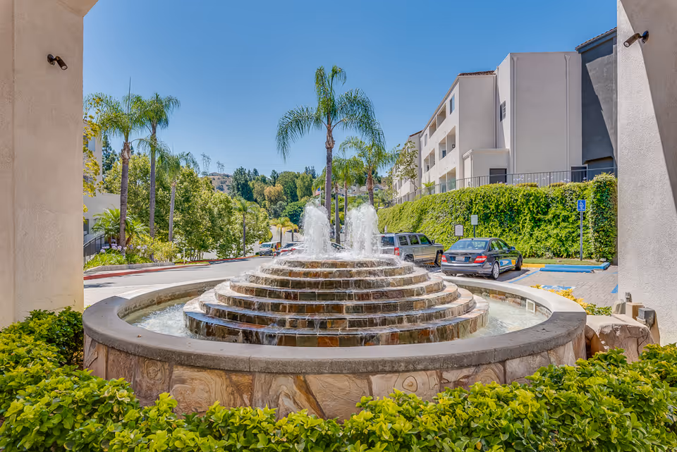 Circular stone fountain centerpiece surrounded by shrubs, palm trees, and a modern senior living building exterior.