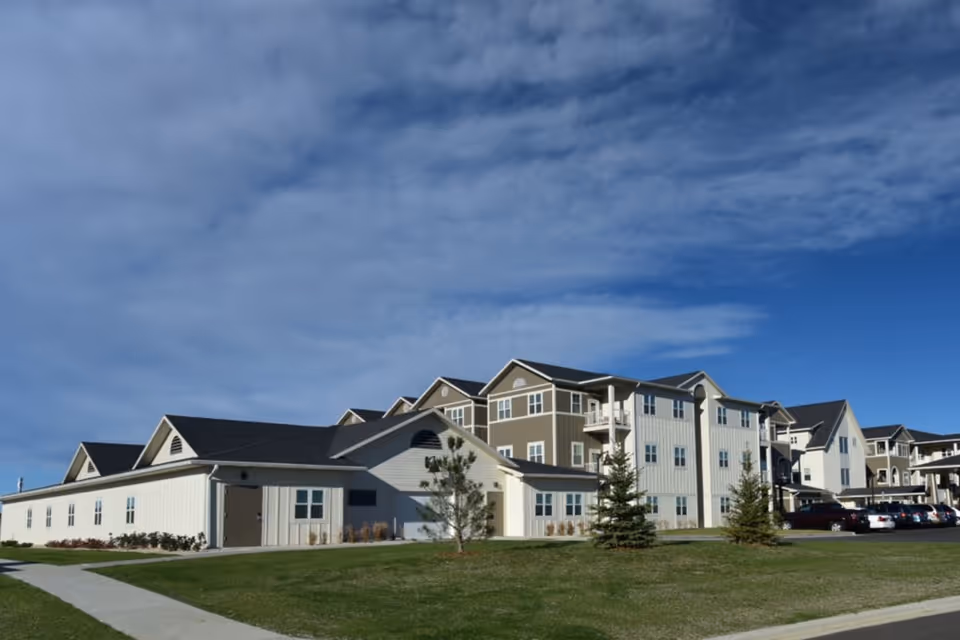 Exterior view of a multi-story senior living facility building with beige and white siding, multiple windows, and a parking area with cars. The sky is partly cloudy with blue patches.