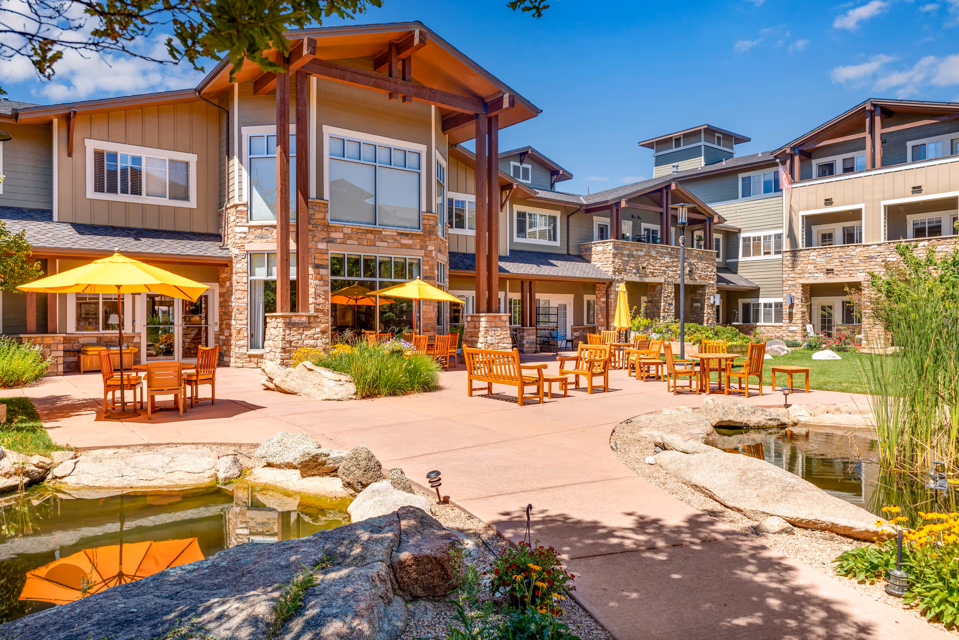 Outdoor patio area at Balfour Louisville Campus featuring wooden tables and chairs with yellow umbrellas, surrounded by a landscaped garden with rocks and a small pond, adjacent to a multi-story building with large windows and stone accents under a clear blue sky.