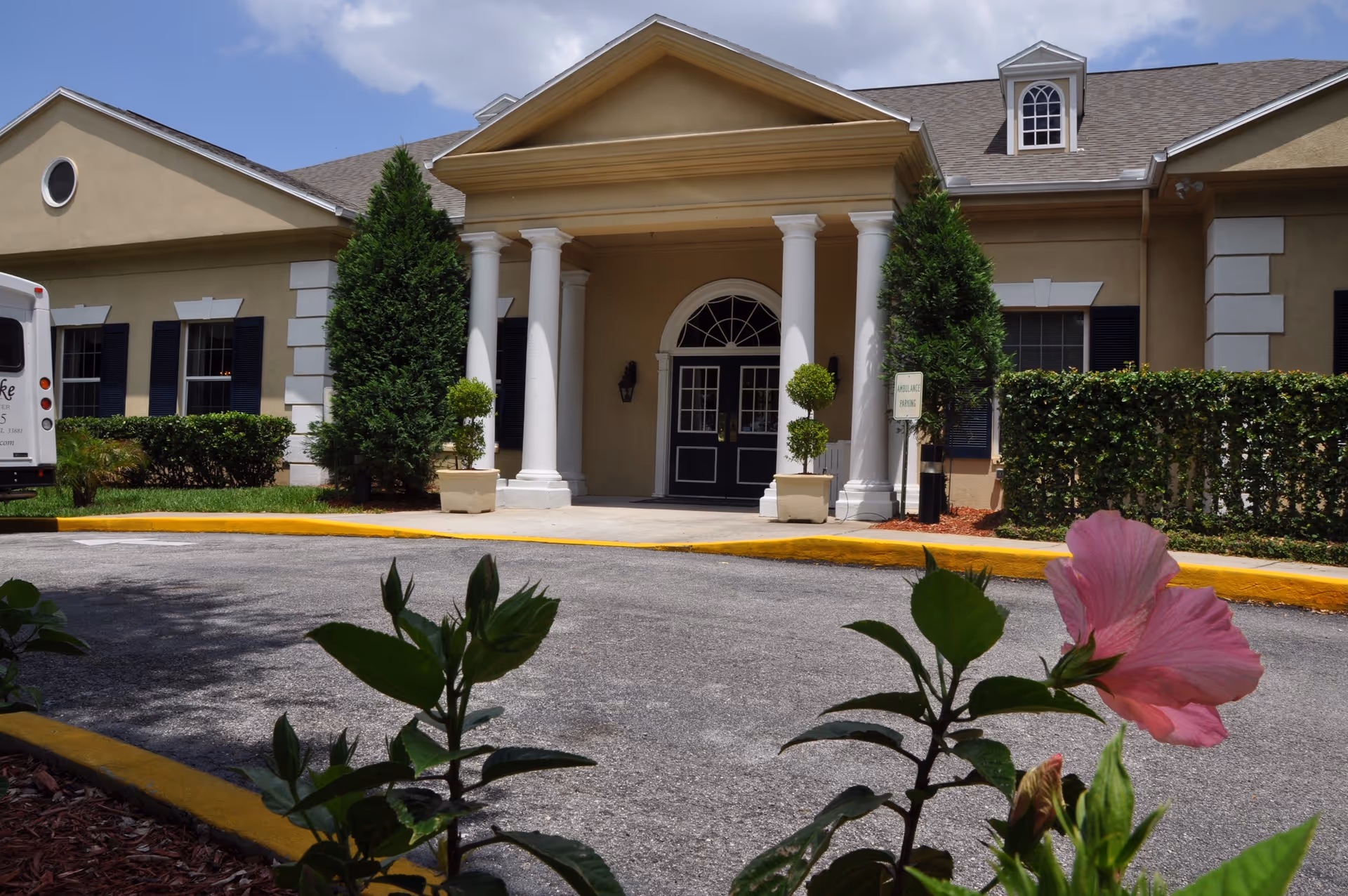 Front view of a beige senior living building with columns, entrance, and landscaped shrubs.