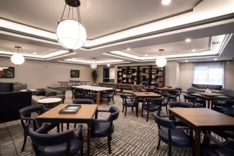 Spacious dining/activity room with multiple tables and chairs, bookshelves along the back wall, and pendant lighting.