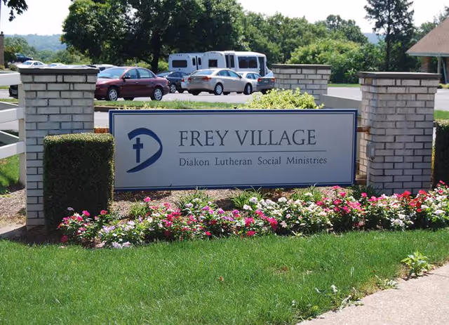 Exterior entrance sign for Frey Village with brick columns, flowers, and a parking lot behind.