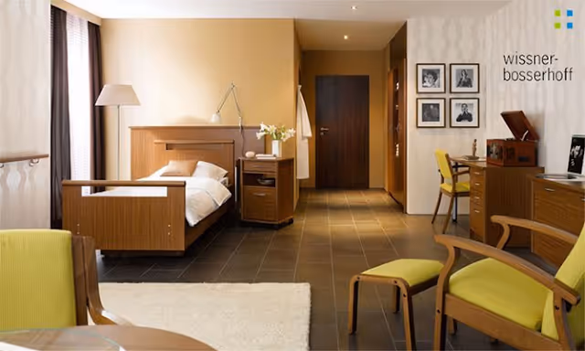 A warm senior living bedroom with a wooden bed, nightstands, desk, and seating area.