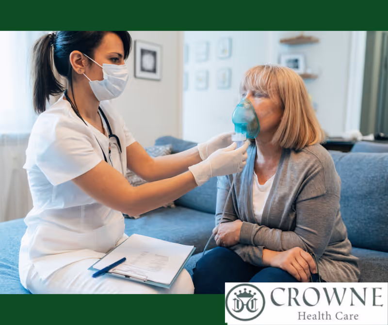 A healthcare professional wearing a mask and gloves is assisting an elderly woman sitting on a couch by helping her use an oxygen mask. The healthcare worker has a clipboard and pen on her lap.