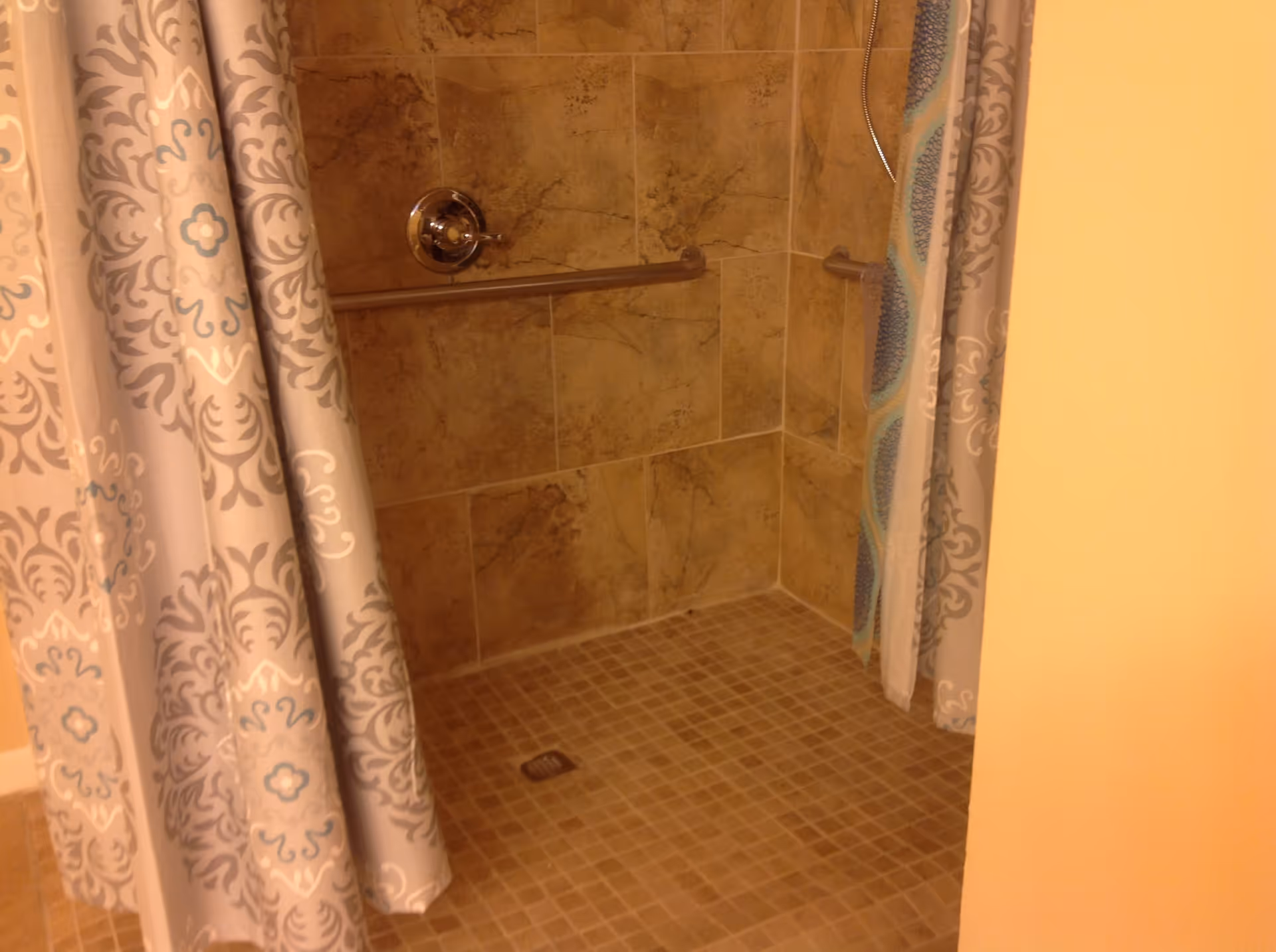 Tiled walk-in shower with patterned curtains, a horizontal grab bar, and a handheld showerhead.