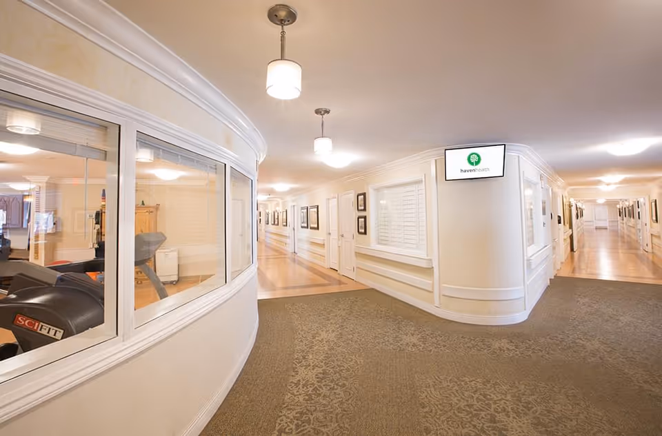 Interior hallway of a senior living facility with beige walls, carpeted and wooden floors, framed pictures on the walls, and ceiling lights. A window on the left shows exercise equipment inside a fitness room. A digital screen on the wall displays the Haven Health logo.