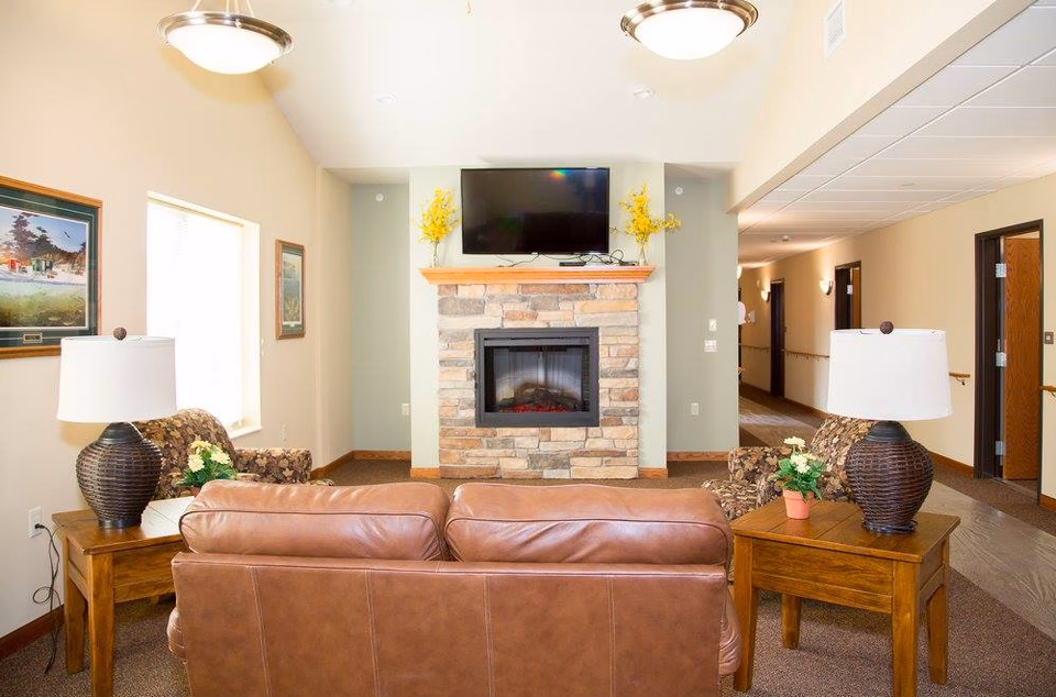 A cozy senior living lounge with a stone fireplace, sofa, chairs, and a mounted TV.
