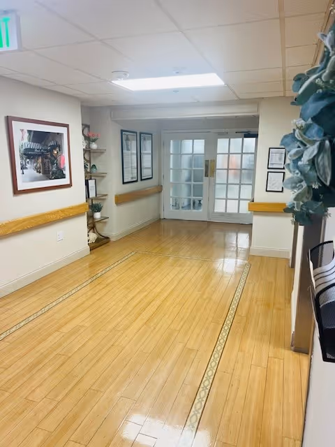 A clean, well-lit hallway with wooden flooring and beige walls. The hallway features handrails on both sides, framed pictures and documents on the walls, a small corner shelf with decorative items, and double glass doors at the end. A green exit sign is visible on the ceiling.
