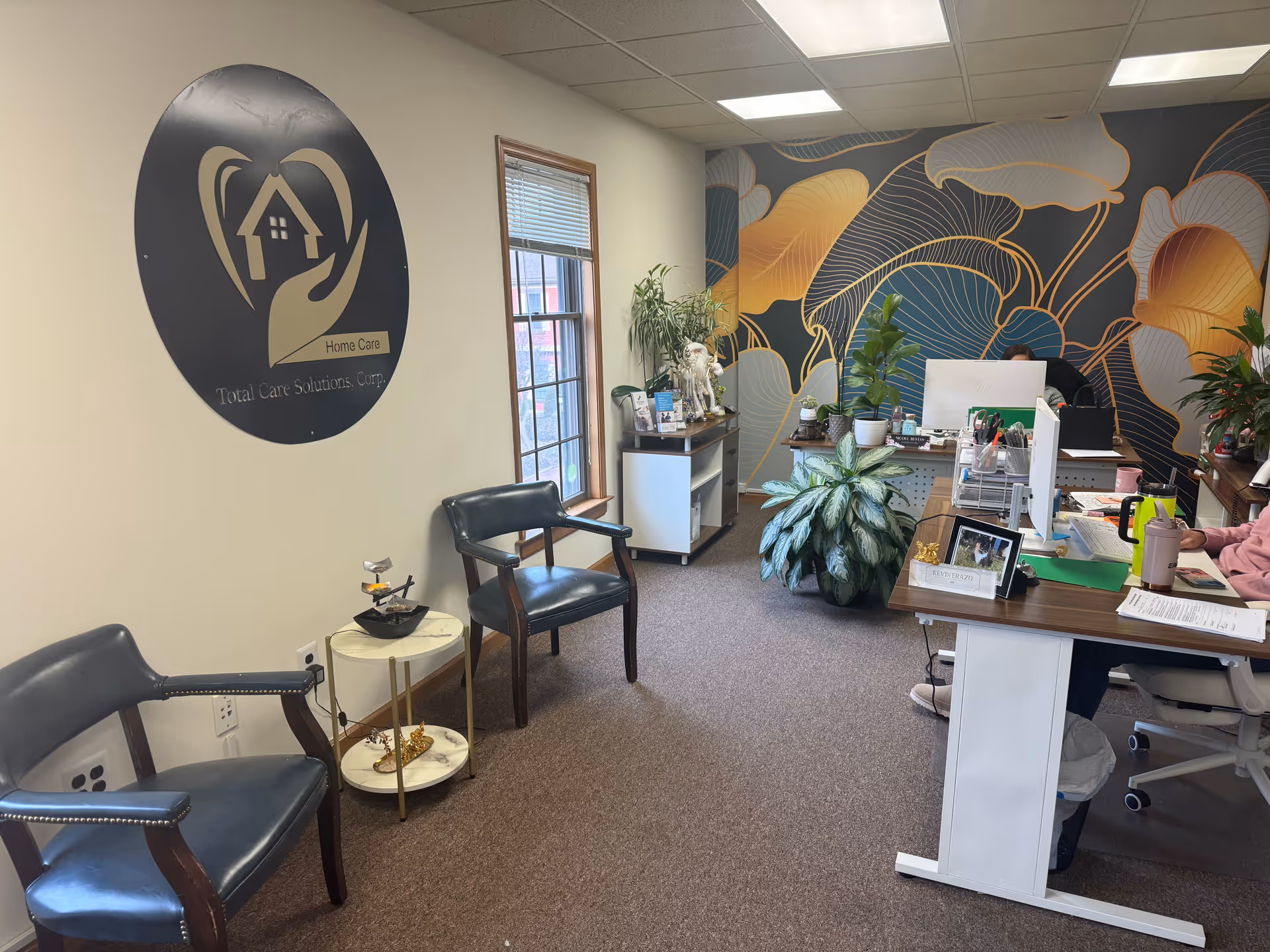 Reception area for Total Care Solutions Corp with logo wall, desks, plants, and seating.