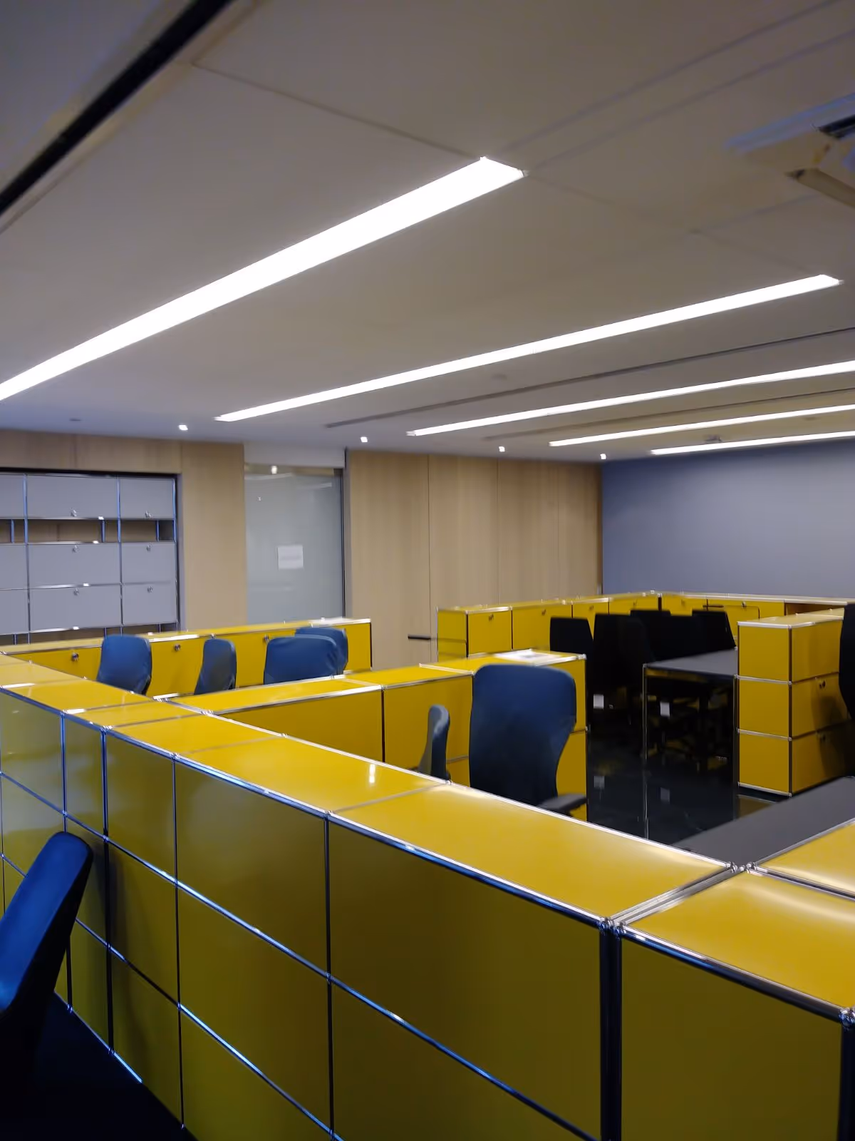 Interior office area with yellow cubicles, blue chairs, and bright overhead lighting.