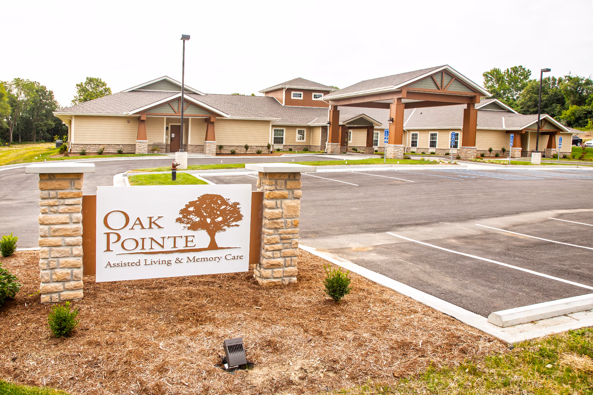Exterior view of Oak Pointe Assisted Living & Memory Care facility showing a large building with a covered entrance and an empty parking lot in front. A sign with the facility name and logo is prominently displayed in the foreground.
