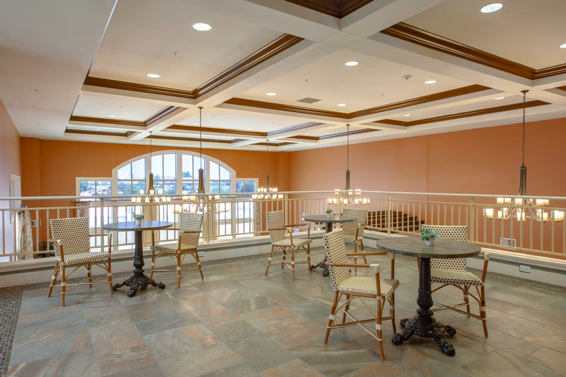 Sunlit interior lounge with round tables, wicker chairs, and a railing overlooking an arched window.