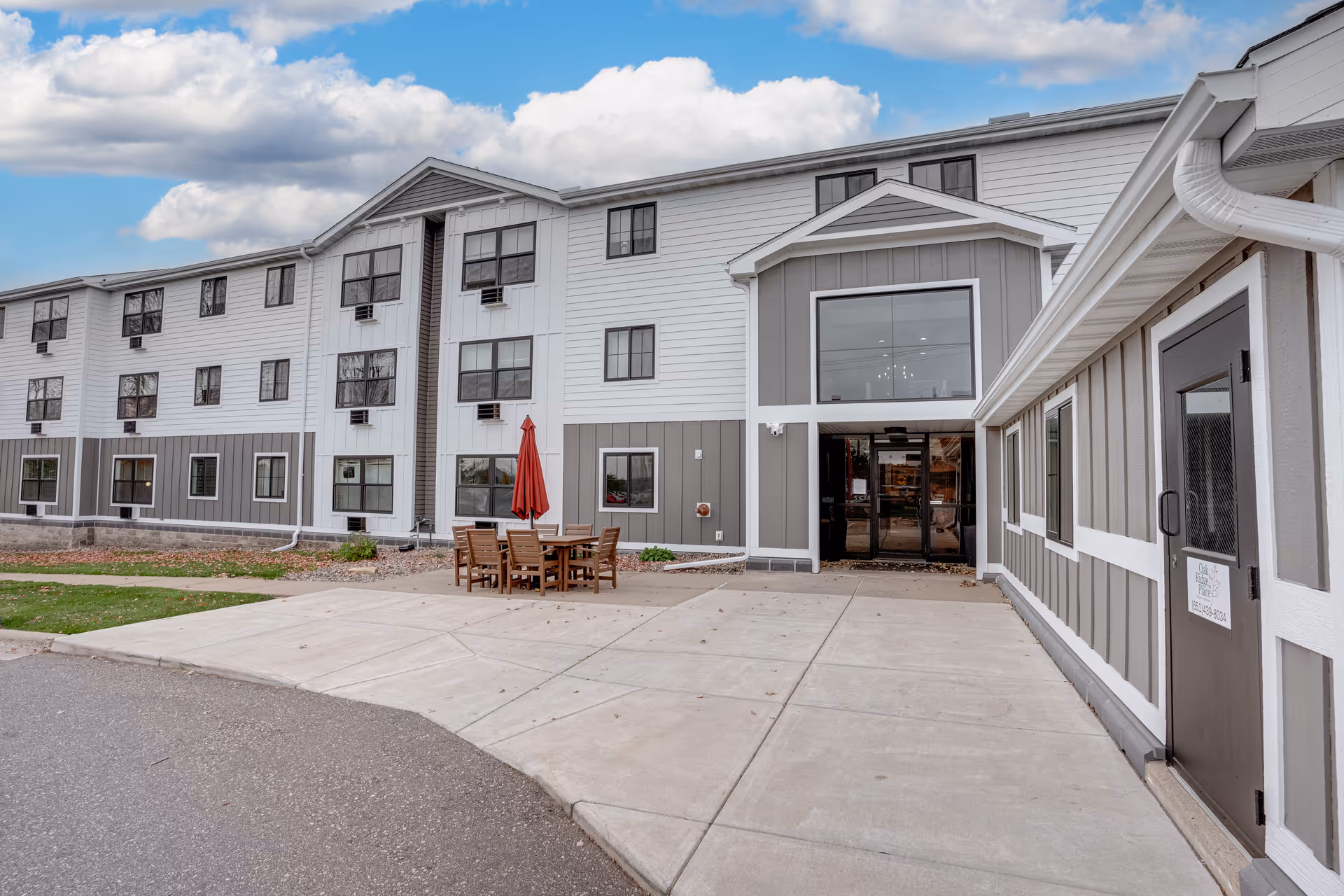 Gray and white senior living building exterior with a patio seating area and red umbrella.