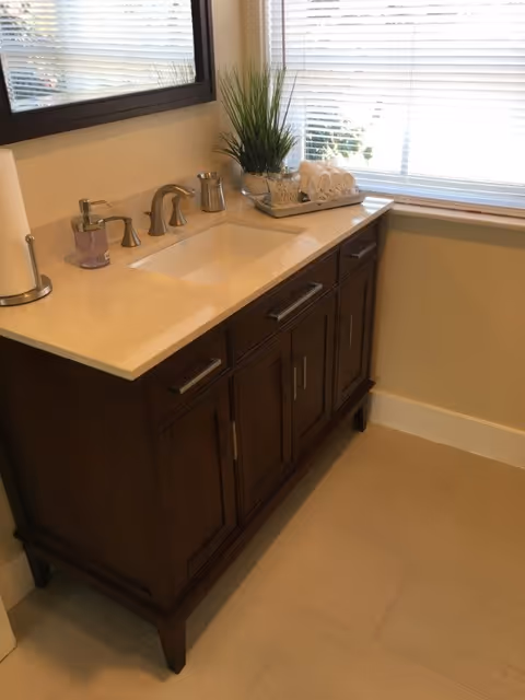 Bathroom vanity with a white countertop sink, chrome faucet, decorative plant and rolled towels beneath a window with blinds.