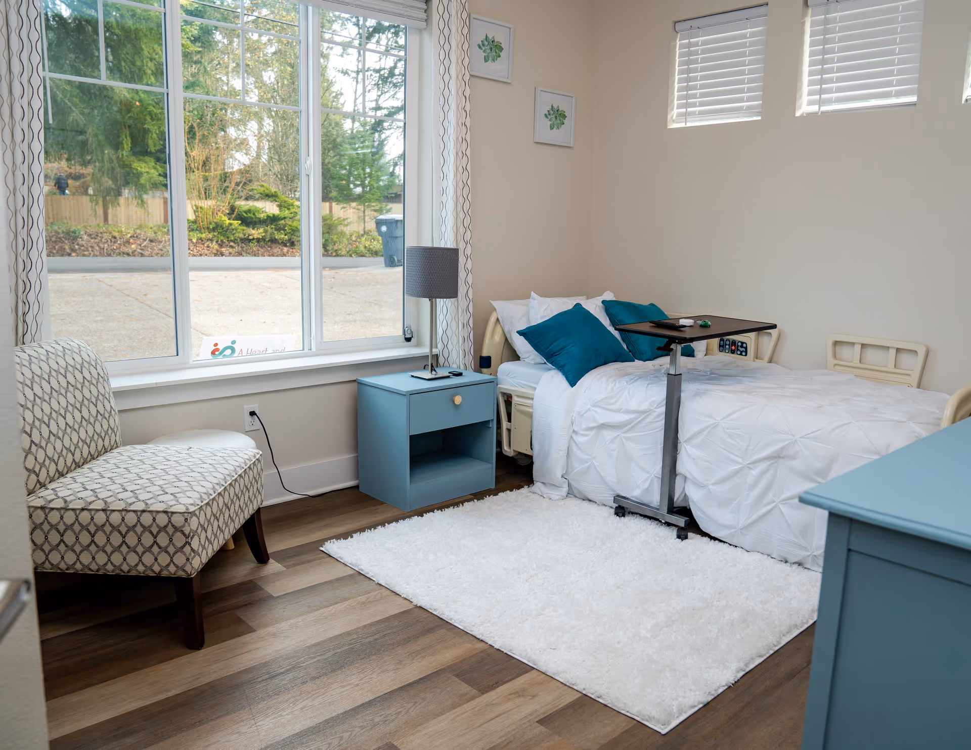 Bright bedroom with a hospital-style bed, blue nightstand, overbed table, and large window.