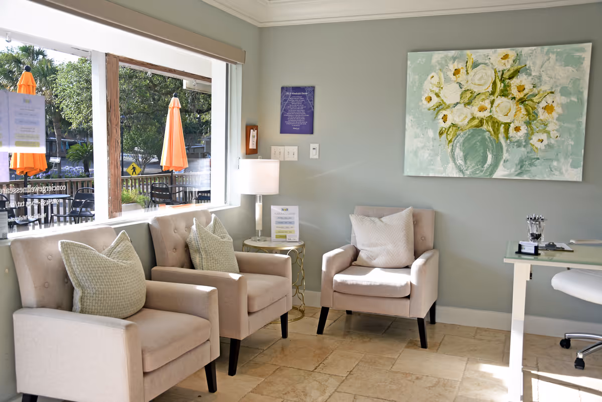 Cozy waiting lounge with beige chairs, a painting, and a large window overlooking outdoor patio.