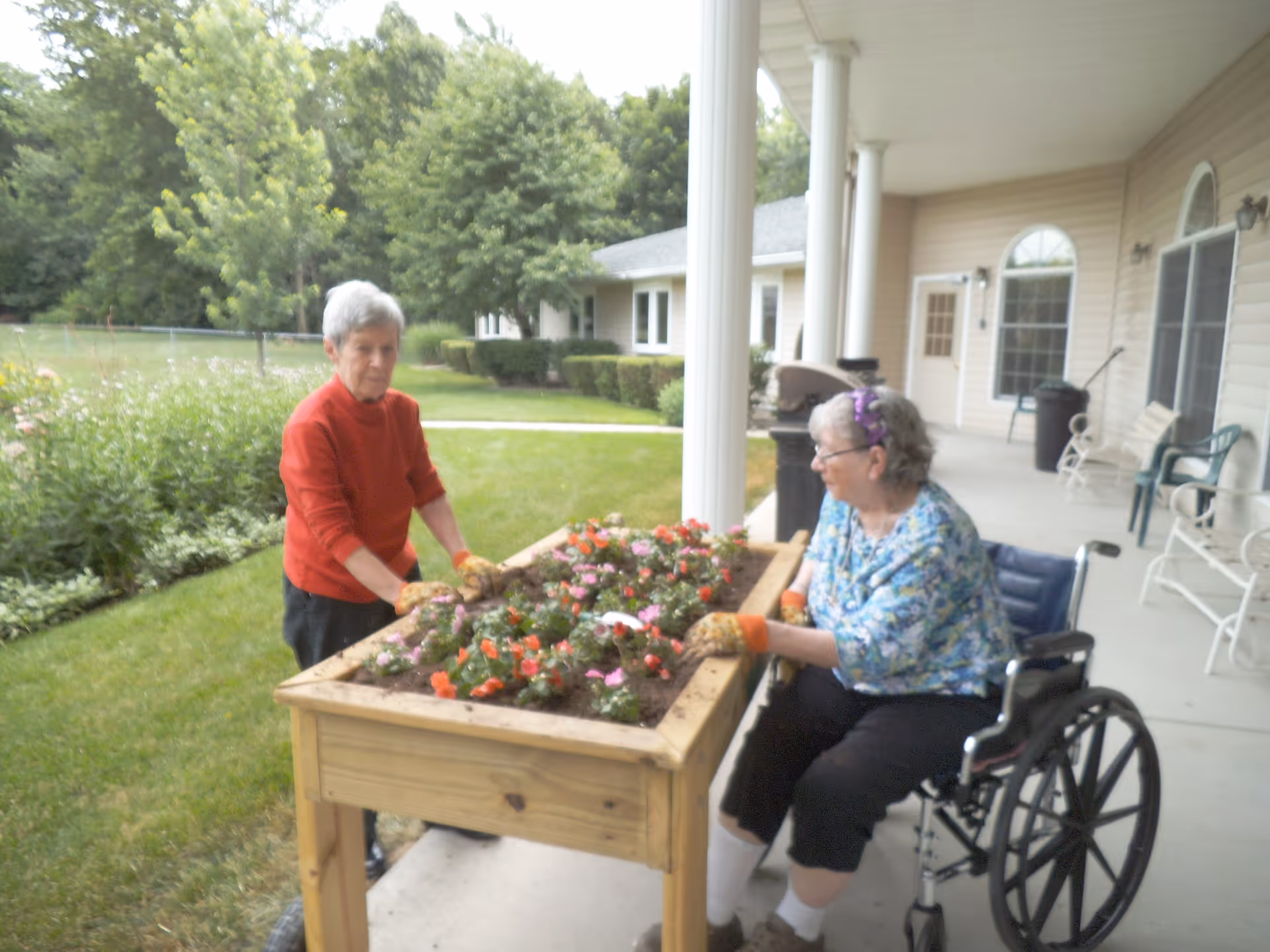 Two older women gardening at a raised wooden planter on a covered patio outside an assisted living building, with one woman in a wheelchair.